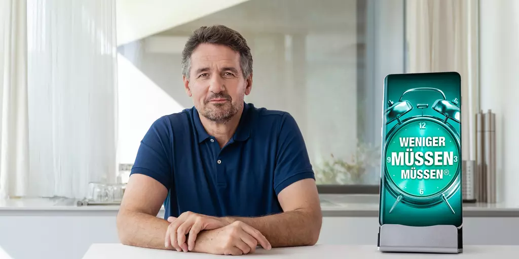 Mann schaut lächelnd in die Kamera, neben ihm steht ein Smartphone mit dem Slogan "Weniger müssen müssen"