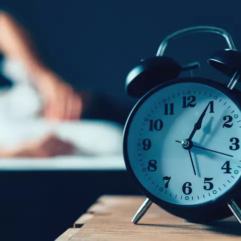 Ein Wecker auf einem Nachttisch, dahinter eine Person, die wach im Bett liegt aufgrund nächtlichem Harndrang
