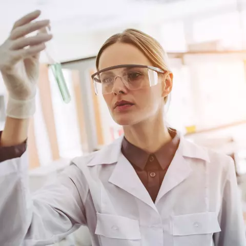 Frau im Labor mit Schutzbrille und weißem Mantel, die eine Probe im Reagenzglas prüft